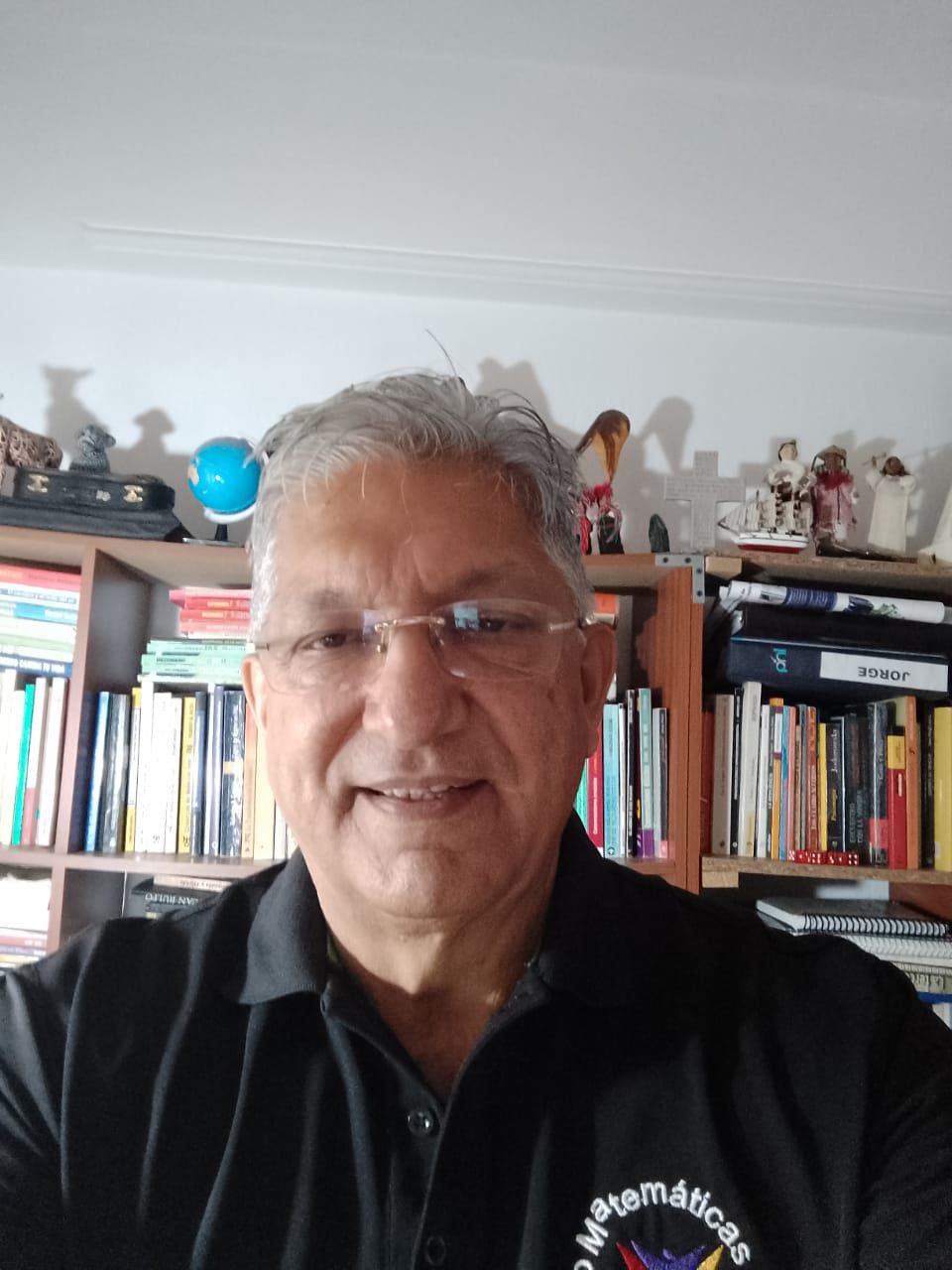 Hombre mayor amigable, de cabello gris, con gafas y un polo negro con el logo de "Matemáticas", en un entorno de oficina en casa.